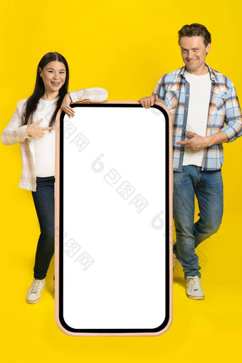 指出白色屏幕高加索人男人。亚洲女人站靠巨大的智能手机空白屏幕移动应用程序广告孤立的黄色的背景产品放置