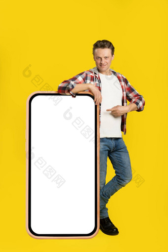 英俊的年轻的男人。站靠巨大的巨大的智能手机数字平板电脑白色黑色的屏幕指出穿着格子衬衫牛仔裤孤立的黄色的背景免费的空间模拟