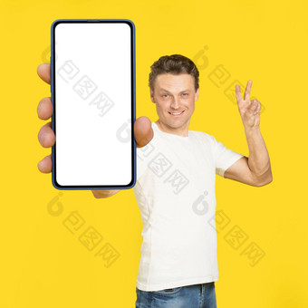 手势胜利英俊的男人。持有巨大的智能手机白色屏幕穿白色t恤牛仔裤孤立的黄色的背景移动应用程序广告伟大的提供