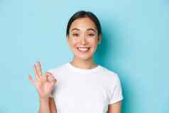 特写镜头漂亮的满意微笑亚洲女孩白色t恤显示建议手势标志点头批准赞美选择同意站光蓝色的背景