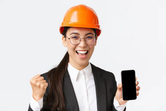 成功的赢得亚洲女工程师架构师安全头盔西装显示智能手机屏幕拳头泵大喊大叫喜乐并成就白色背景