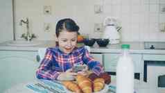 美丽的女孩玩智能手机早....坐着表格厨房童年人技术概念