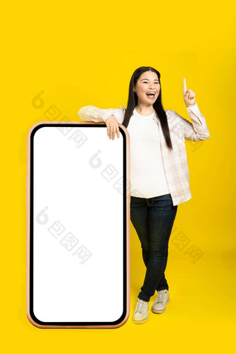 中间年龄亚洲女人兴奋指出手指靠巨大的巨大的智能手机空白白色屏幕穿休闲装孤立的黄色的背景免费的空间模拟
