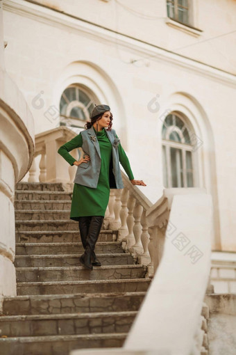 户外时尚肖像优雅的时尚浅黑肤色的女人模型贝雷帽绿色衣服灰色的马甲摆姿势日落欧洲城市秋天