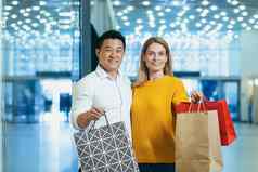 年轻的多样化的家庭亚洲男人。金发女郎女人购物超市肖像夫妇购物者相机微笑持有色彩斑斓的袋货物购物