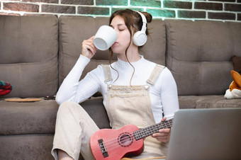 舒适的年轻的赶时髦的人女人休闲穿饮料热咖啡白色杯需要打破学习玩尤克里里琴吉他坐着地板上生活房间首页休闲概念