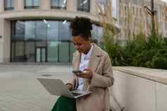 年轻的非洲美国女人学生工作在线笔记本电脑移动电话背景城市现代建筑