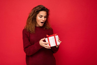 有吸引力的积极的惊讶年轻的浅黑肤色的女人卷曲的女人孤立的红色的背景墙穿白色毛衣持有礼物盒子unboxing现在盒子红色的丝带