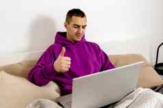 年轻的高加索人男人。紫罗兰色的连帽衫坐着床上持有移动PC电脑男人。笔记本研究在线调用朋友距离学习videocall积极的标志