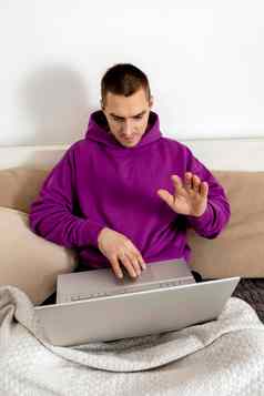 年轻的高加索人男人。紫罗兰色的连帽衫坐着床上持有移动PC电脑男人。笔记本研究在线调用朋友距离学习videocall放松首页