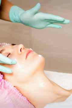 美容师戴着手套手适用于保湿面具剥奶油女脸面部美容治疗程序面部护理