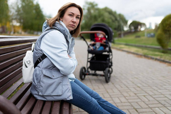 生气年轻的妈妈。坐着公园板凳上婴儿巡回演出的演员愤怒的脸概念产后抑郁症