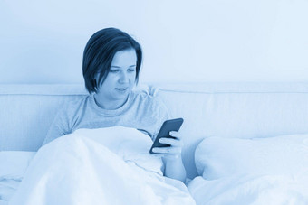 女人智能手机坐在床上覆盖羽绒被