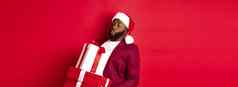 圣诞节一年购物概念有趣的非洲美国男人。圣诞老人他携带重圣诞节礼物持有礼物站红色的背景