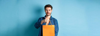英俊的有胡子的男人。拉伸手橙色购物袋使礼物站蓝色的背景