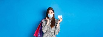 科维德流感大流行生活方式概念时尚的女人穿医疗面具购物预防测量冠状病毒显示塑料信贷卡持有袋蓝色的背景