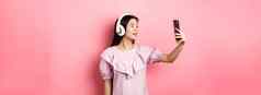 美丽的亚洲女孩博主采取自拍无线耳机使照片社会媒体智能手机站衣服粉红色的背景