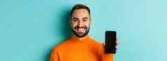 特写镜头英俊的有胡子的的家伙橙色毛衣显示智能手机屏幕微笑显示促销在线绿松石背景