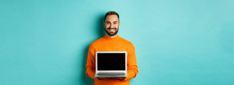 英俊的有胡子的男人。橙色毛衣显示移动PC屏幕展示在线商店站光蓝色的背景