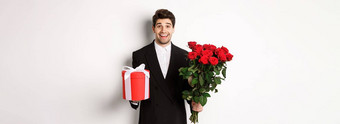 概念假期的关系庆祝活动图像英俊的微笑的家伙黑色的西装持有花束红色的玫瑰给现在一年白色背景