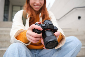微笑红色头发的人女孩摄影师检查照片持有相机屏幕需要照片在户外走街streetstyle<strong>拍摄</strong>