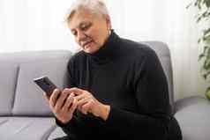 快乐微笑老女人电话屏幕坐着舒适的沙发生活房间兴奋成熟的高级女聊天社会网络购物在线有趣的智能手机