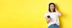 很酷的亚洲女孩跳舞听音乐无线耳机持有智能手机手站黄色的背景