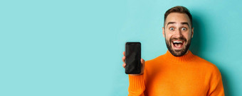<strong>特写</strong>镜头英俊的有胡子的的家伙橙色毛衣显示智能手机屏幕微笑显示促销在线绿<strong>松石</strong>背景
