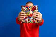 肖像快乐的男人。红色的毛衣持有礼物盒子前面房子孤立的蓝色的背景惊喜概念假期幸福情绪面部表达式庆祝活动