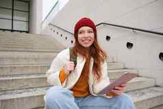 年轻的美丽的女孩长红色的头发微笑互联网应用程序连接无线网络校园坐在楼梯数字平板电脑
