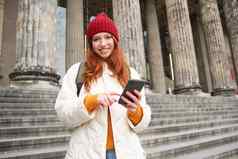 肖像年轻的红色头发的人女人持有移动电话智能手机应用程序走城市中心地图应用程序发短信消息