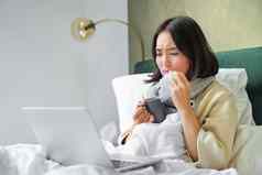 肖像亚洲女孩打喷嚏擦拭流鼻涕的鼻子感觉生病的住床上流感移动PC
