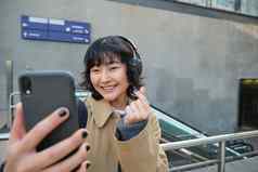 可爱的时尚的朝鲜文女孩穿耳机需要自拍智能手机旅游记录视频使照片站街