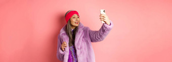 时尚概念美丽的亚洲高级夫人采取自拍智能手机摆姿势聚会，派对闪闪发光的衣服紫色的人造外套站粉红色的背景