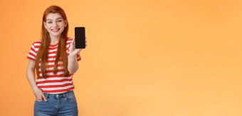 快乐的好看的红色头发的人女人<strong>介绍</strong>社会媒体<strong>功能产品</strong>持有智能手机显示相机手机显示促进设备应用程序微笑广泛的橙色背景