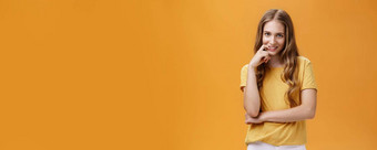 有创意的棘手的年轻的女人自然波浪长头发黄色的t恤额头意图欲望表达式咬手指微笑相机橙色墙