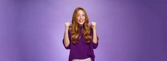 是的使快乐快乐的年轻的漂亮的红色头发的人女孩紫色的毛衣紫罗兰色的背景提高握紧的拳头快乐胜利庆祝成功赢得欢呼