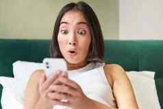 可爱的亚洲女孩持有智能手机惊讶移动电话屏幕阅读消息惊讶脸表达式