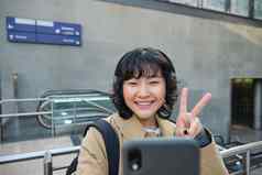 可爱的时尚的朝鲜文女孩穿耳机需要自拍智能手机旅游记录视频使照片站街