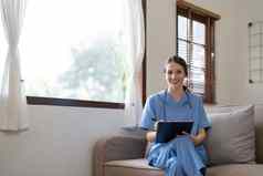 肖像年轻的有吸引力的微笑护士穿统一的听诊器坐着沙发首页护士高加索人女人工作护理首页