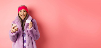 在线购物时尚概念快乐亚洲女检查互联网促销活动持有信贷卡智能手机粉红色的背景