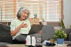 上了年纪的女人坐着沙发上在线咨询医生移动PC远程医疗咨询远程医学概念