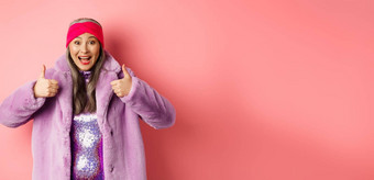 时尚<strong>购</strong>物概念快乐的亚洲女人显示竖起大拇指批准推荐促销活动站时尚的<strong>紫色</strong>的皮毛外套粉红色的背景