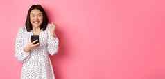 在线购物美概念兴奋亚洲女人赢得互联网持有智能手机欢乐微笑快乐庆祝站粉红色的背景