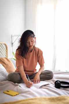 亚洲女青少年大学学生坐着床上写作杂志舒适的卧室复制空间垂直图像