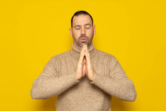 有胡子的拉美裔男人。穿米色高领毛衣孤立的黄色的背景持有手祈祷冥想放松平静做梦希望