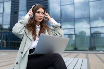 年轻的女人耳机移动PC膝盖听音乐背景玻璃办公室外观