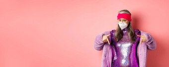 冠状病毒购物概念时尚的亚洲上了年纪的女人紫色的衣服脸面具指出手指显示促销粉红色的背景