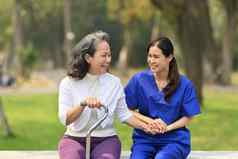 微笑亚洲女护士持有高级女人手愉快的谈话坐着户外康复中心