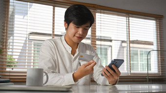 英俊的成人亚洲男人。消费者持有信贷卡智能手机购物在线在线银行事务